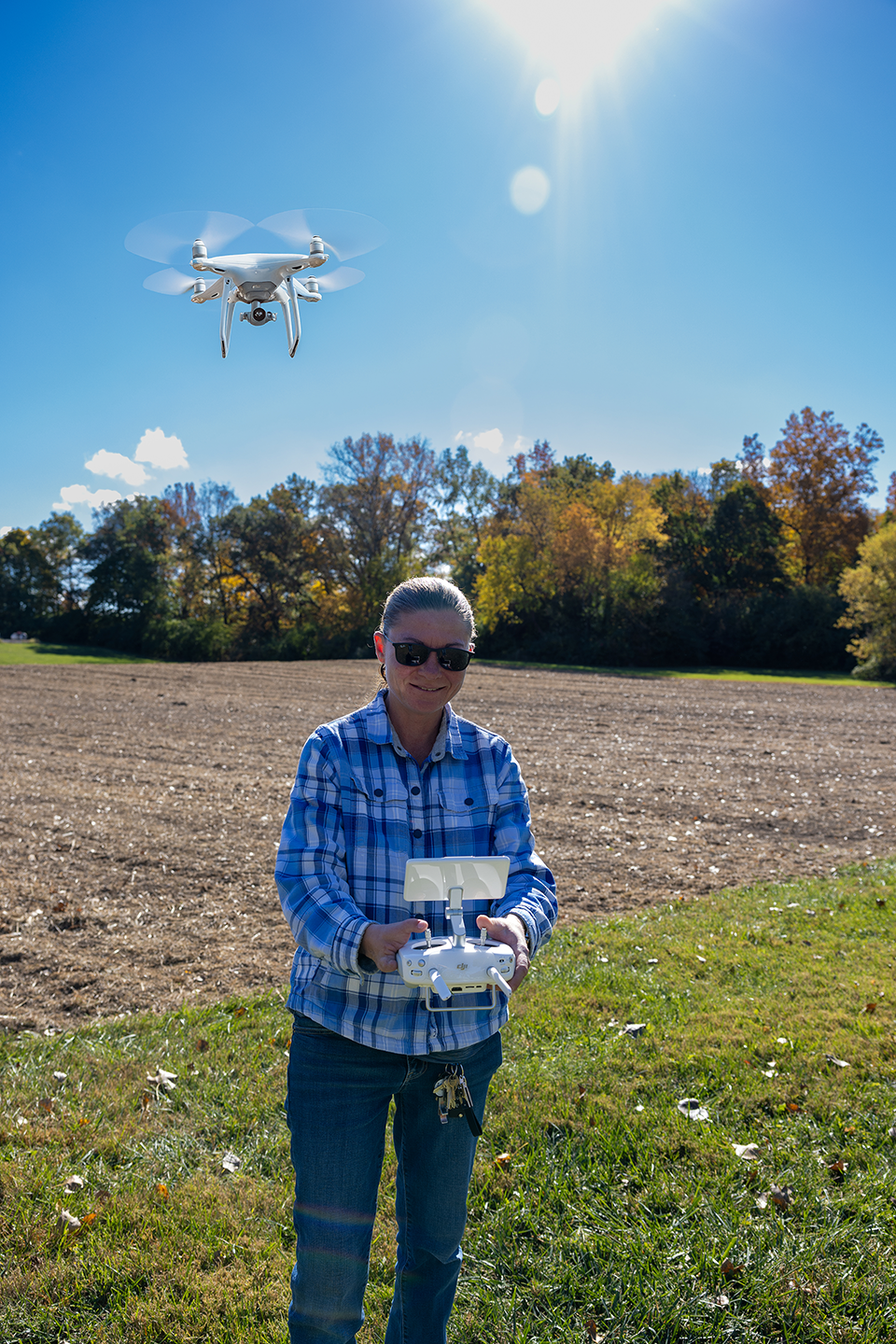 Woman standing in agriculture field holding a remote control for a drone flying in the background. 