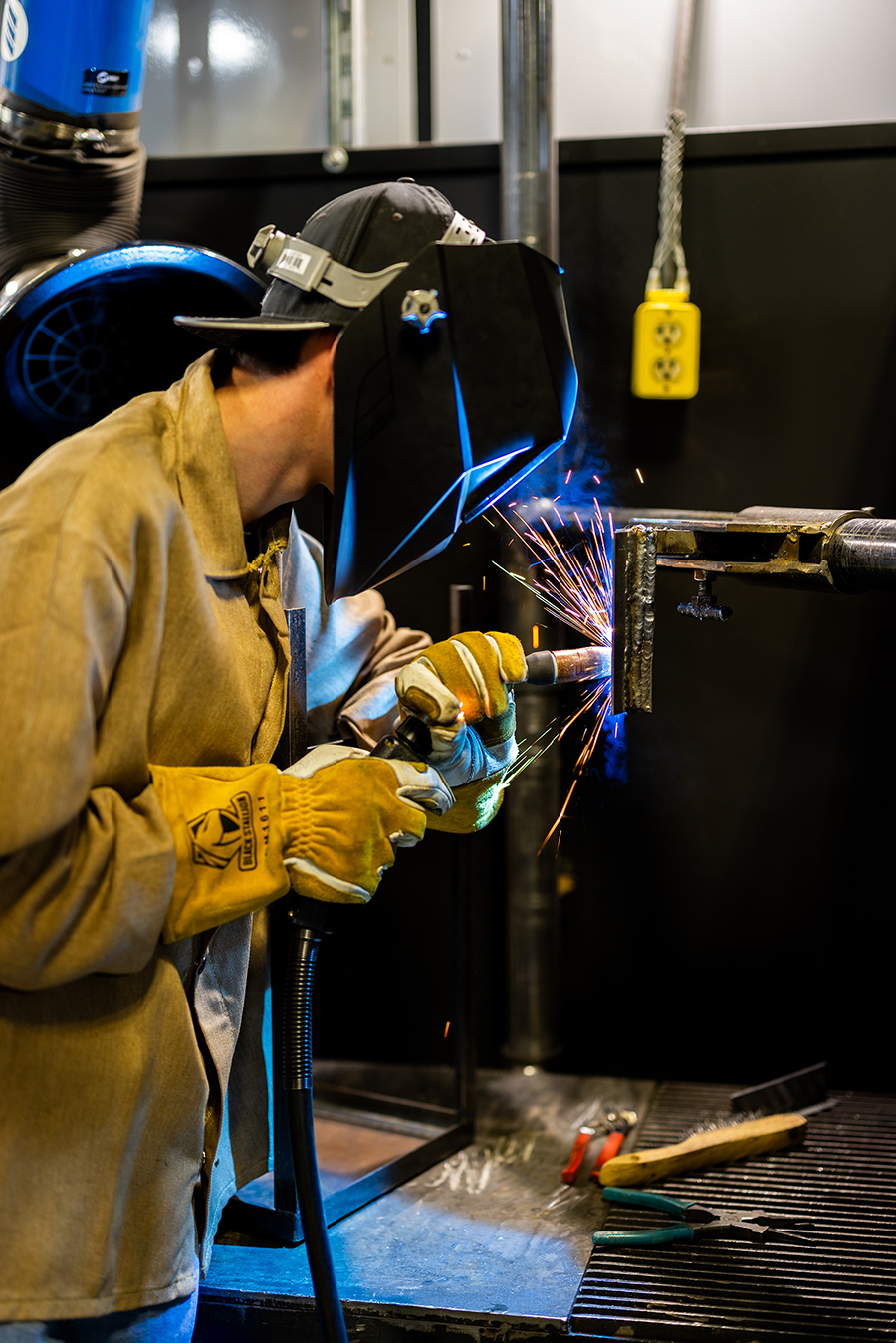 Man welding with sparks flying, wearing yellow safety coat, yellow gloves and a welding helmet.