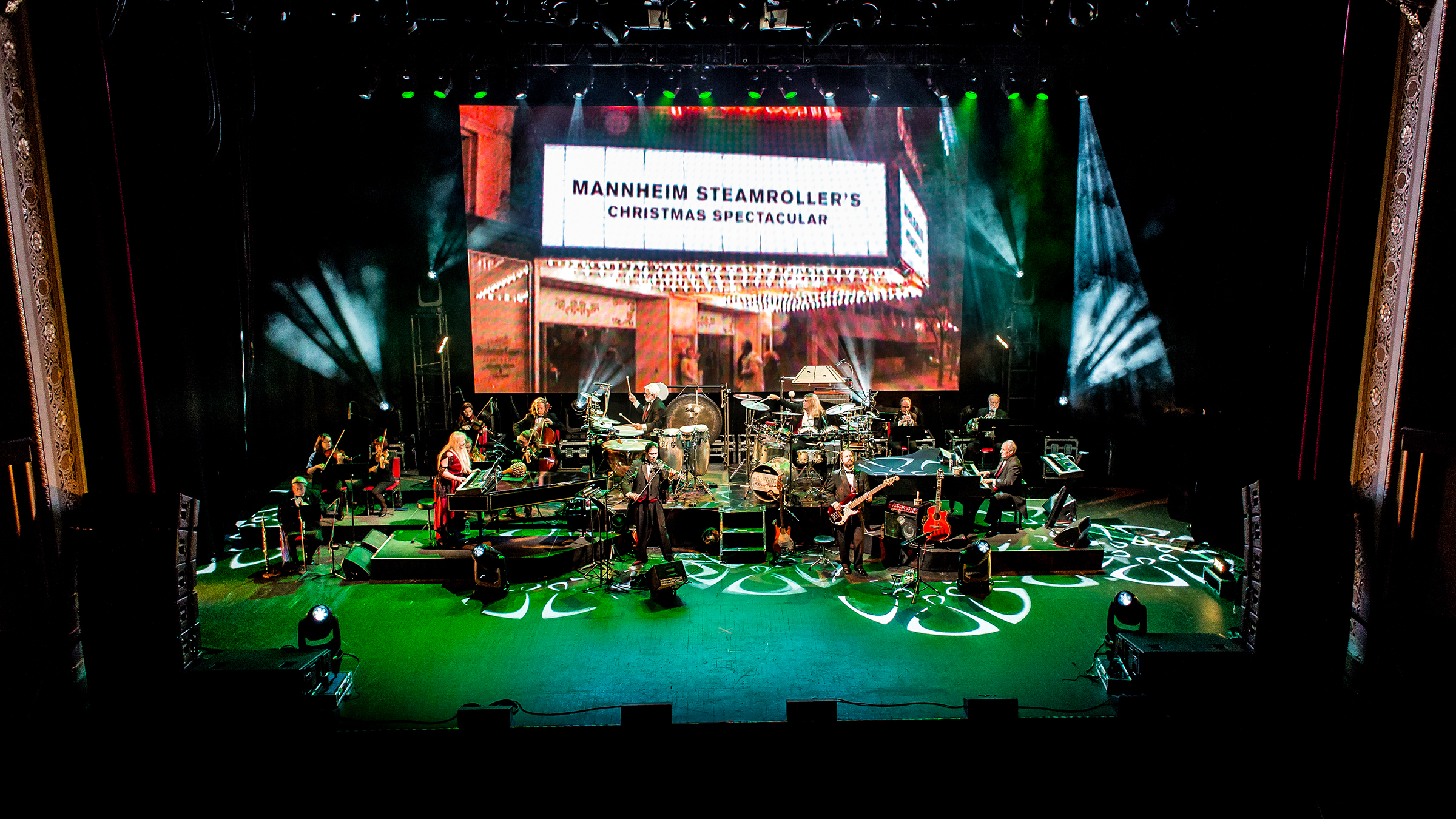 Concert stage bathed in green light with musicians and Mannheim Steamroller backdrop.