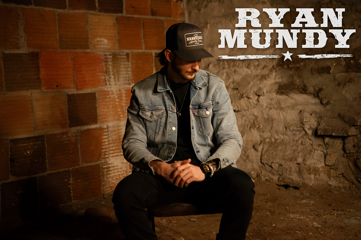 Rock Country artist Ryan Mundy sitting against brick wall wearing light blue jean jacket and brown ball cap.