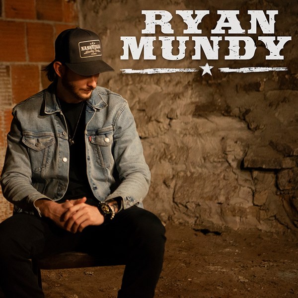 Rock Country artist Ryan Mundy sitting against brick wall wearing light blue jean jacket and brown ball cap.