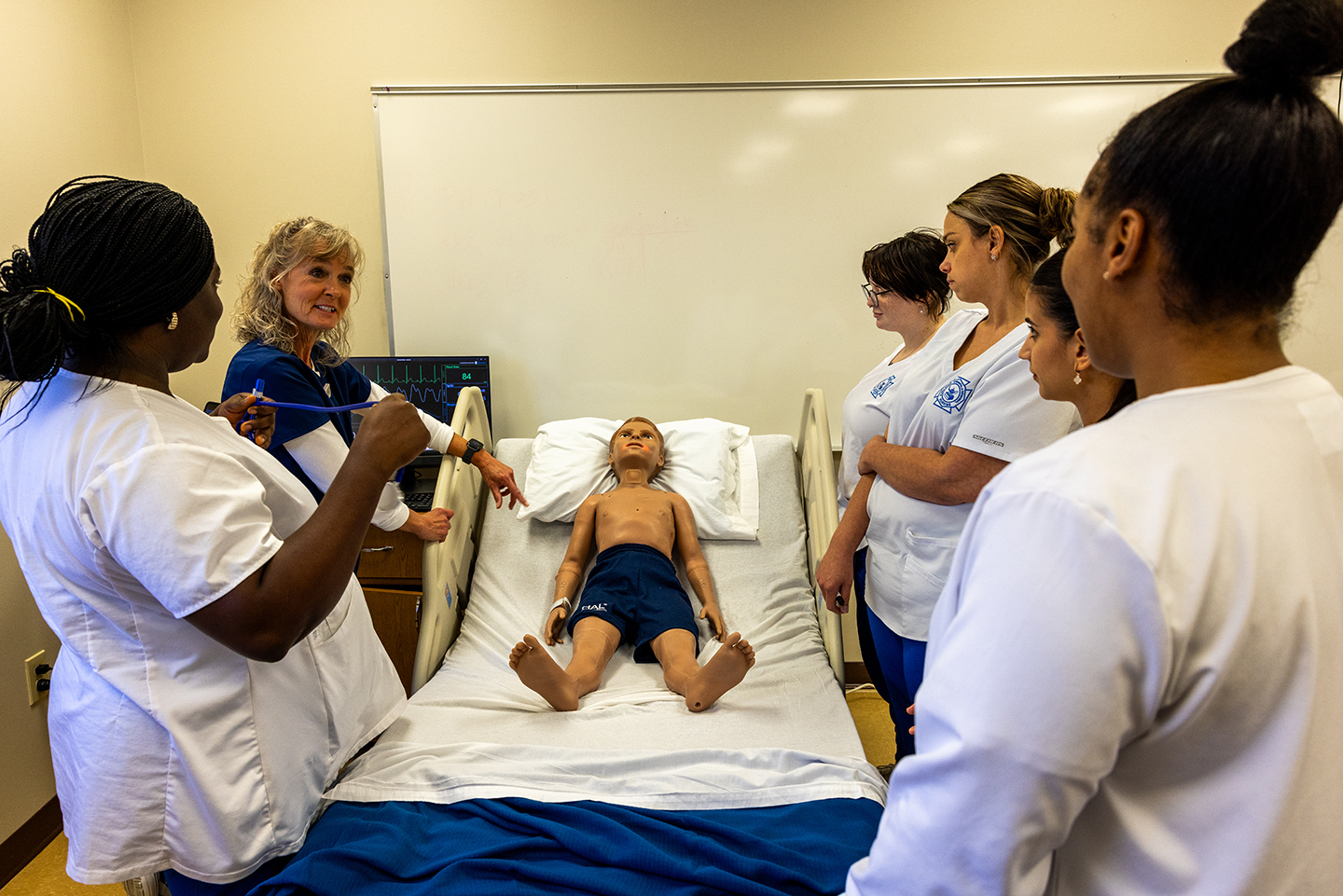 Nursing instructor with blonde hair with five female nursing students and mannequin patient.