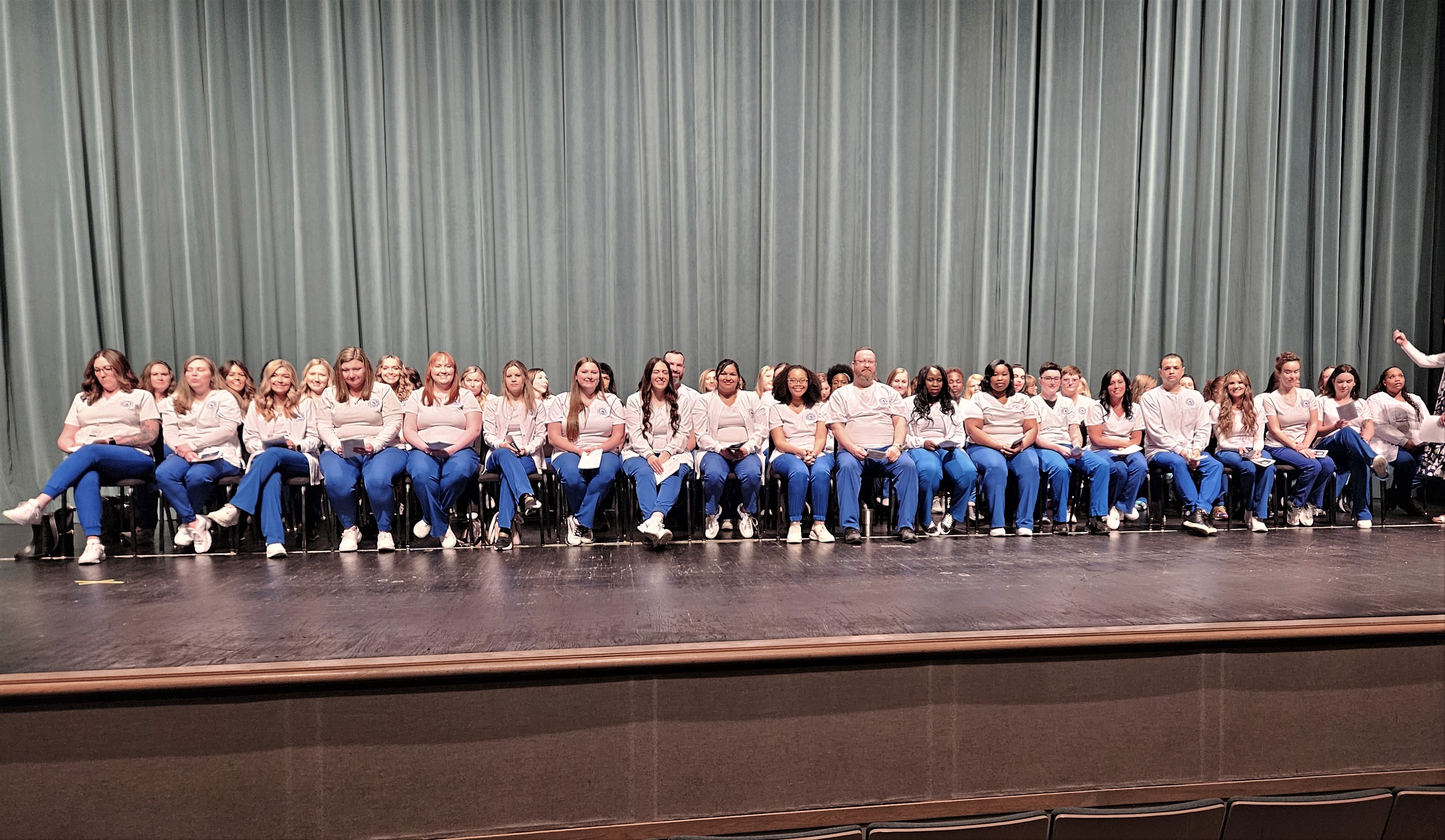 Kuss Auditorium stage with 78 nursing students sitting in chairs wearing blue scrub pants and white shirts 