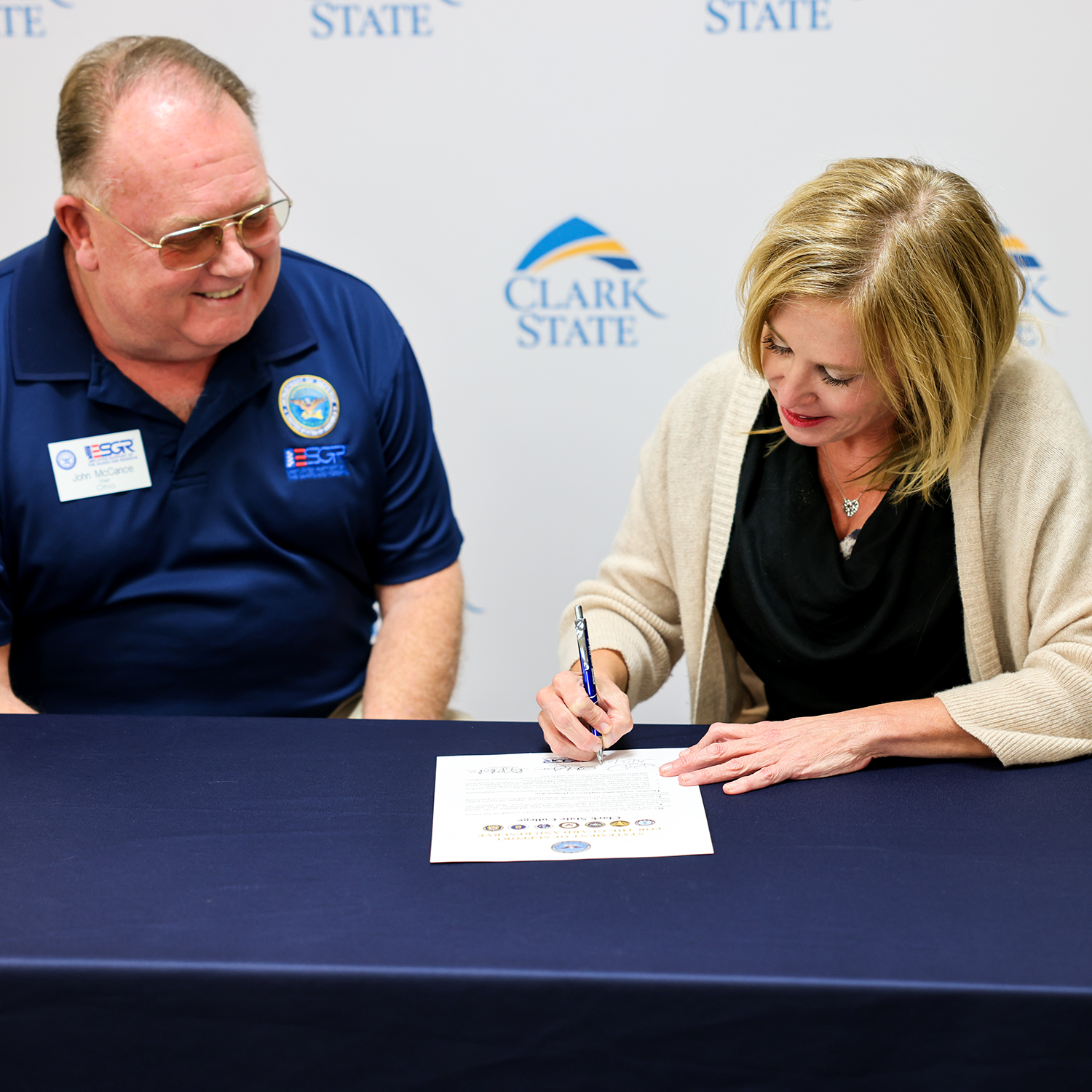 Man on left in blue shirt; woman on right in cream sweater and blonde hair signing commitment paper.